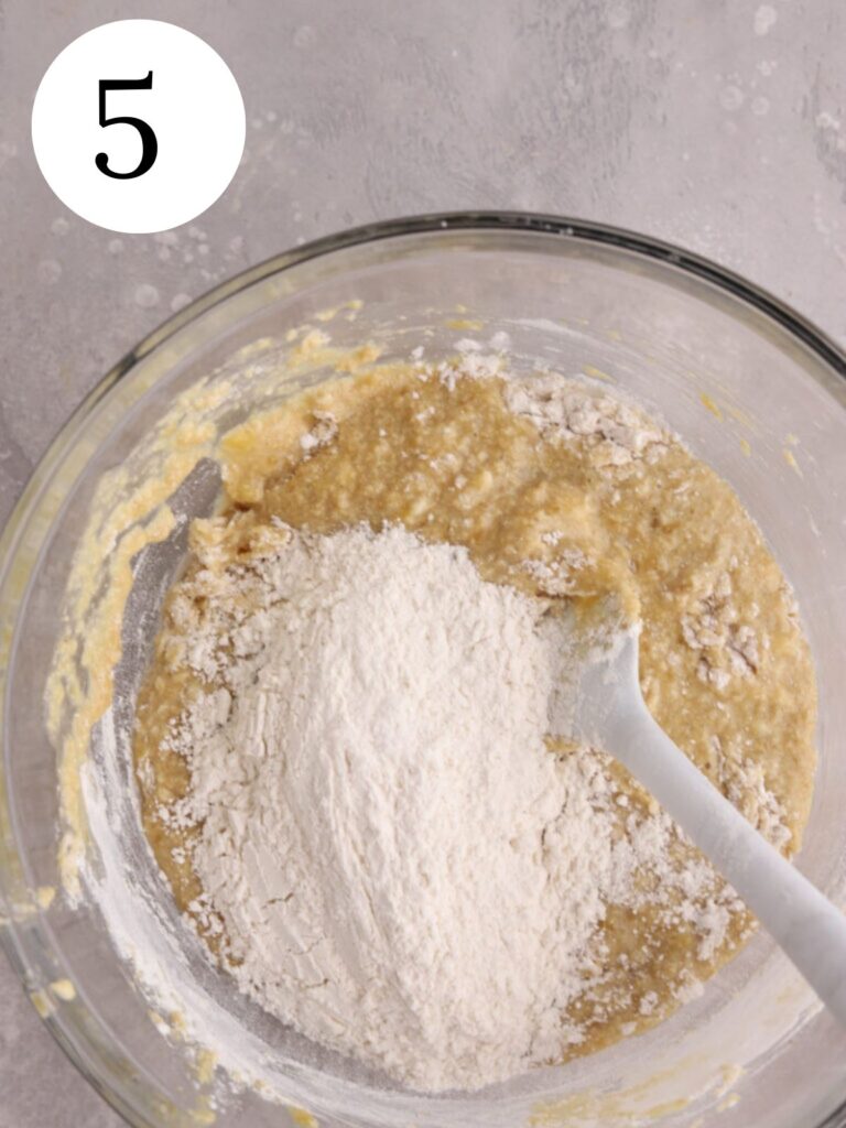 A mixing bowl with flour being stirred into batter with a spatula.