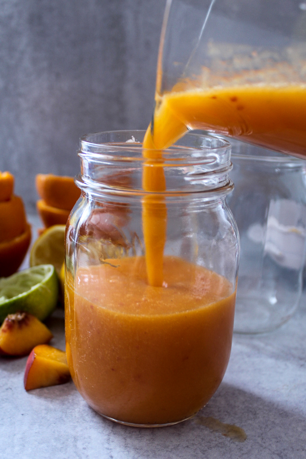 a mason jar with fresh orange peach mango smoothie getting poured into it and an empty mason jar behind it with some peach slices, and juiced lime, lemon and orange peels behind it 