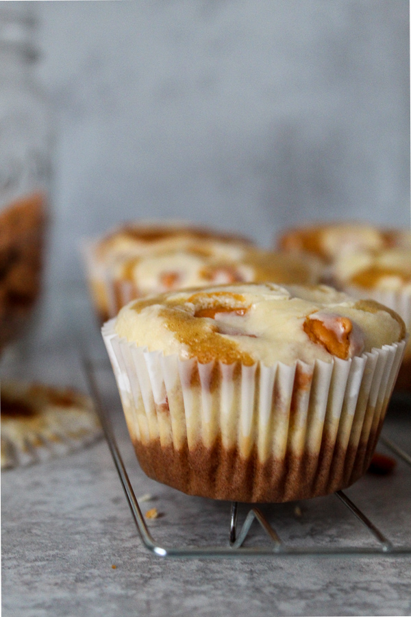 A closeup of a butterscotch and cream cheese cupcake with a butterscotch chip popping out on top and half covered with baked cream cheese on a cooling rack with other cupcakes out of focus behind it and a jar of butterscotch chips in the background 