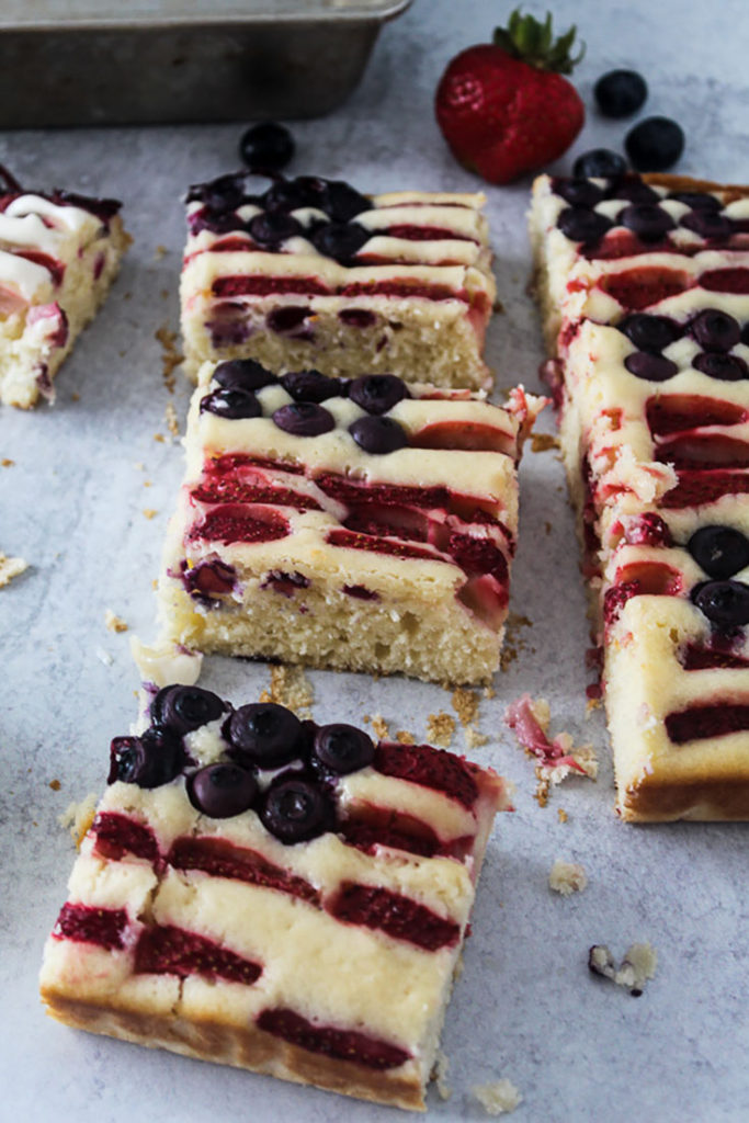 Square slices of pieces of american flag cake decorated with blueberries and strawberry slices on a counter with a strawberry and three blueberries and a cake pan behind the cake 