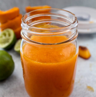 a mason jar full of a bright orange mango orange lemon lime smoothie with a couple of drops of the smoothie on the counter next to it and an orange napkin in the front corner of the picture. Behind the smoothie are the peels of juiced oranges stacked on top of each other, juiced limes and a peach slice and a blender top
