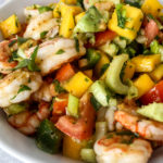 a large white bowl of shrimp salad with avocado and mango and a lemon next to it
