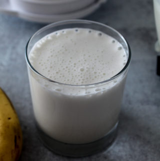 a glass full of a frothy, creamy banana smoothie with a blender lid behind it and a banana next to it
