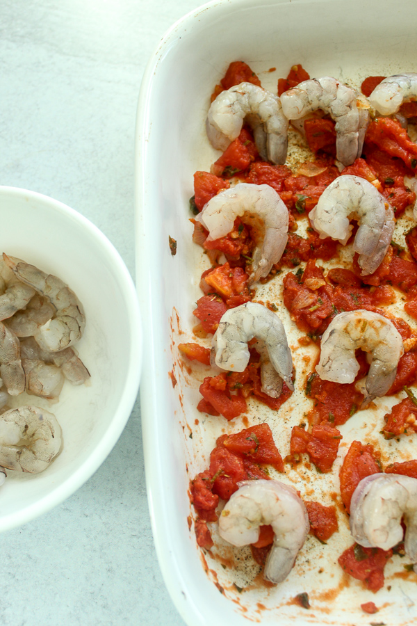 an overhead image of a baking tray with tomato sauce on the bottom of it and rows of uncooked shrimp on it and a bowl of uncooked shrimp next to it