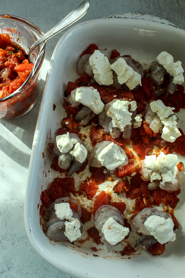 a white baking dish with tomato sauce on the bottom of it and rows of shrimp topped with goat cheese and a glass measuring cup on the side full of tomato sauce with a spoon in it