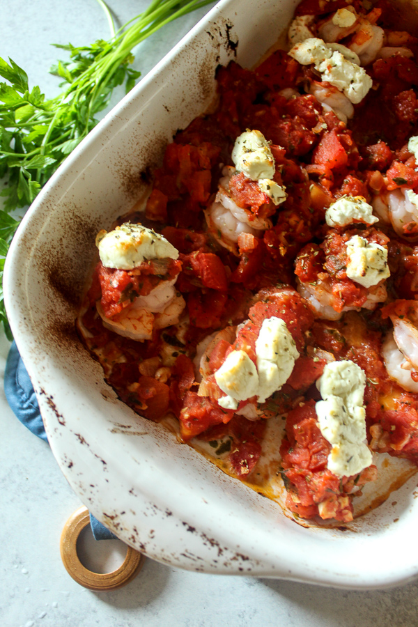 a white baking dish with baked shrimp covered with tomato sauce, goat cheese, and herbs on a blue oven mitt and a bunch of parsley beside it