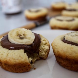 nutella filled sugar cookie cups with a banana on top on a white plate with a knife and jar of nutella in the background