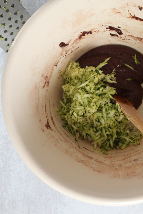 a tan mixing bowl with chocolate batter and shredded zucchini in it and a wooden spoon 