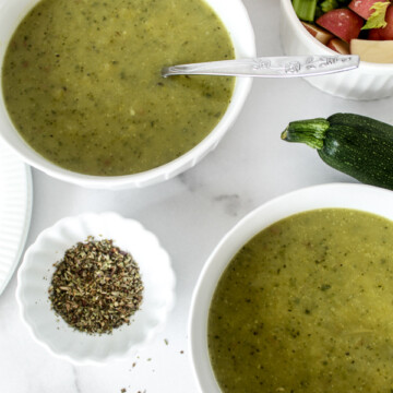 an overhead view of two white bowls of green creamy zucchini soup with a small bowl of dried herbs next to it and a bowl of chopped vegetables and a zucchini next to it