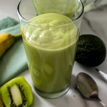 a creamy green smoothie in a class with a kiwi peeled and sliced next to it and a banana on the counter and a blender behind it and an avocado next to it