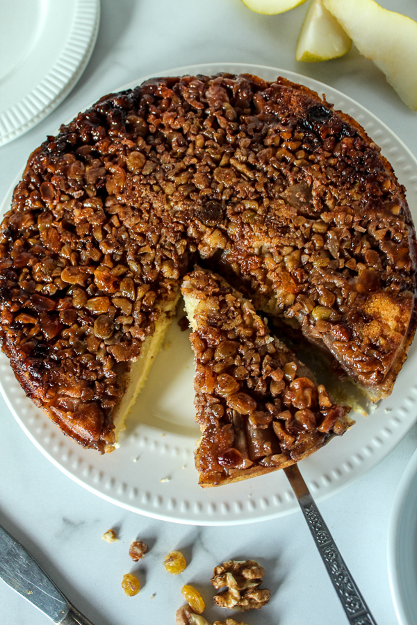 an overhead view of upside down pear and peach cake on a white plate with raisins and walnuts on the counter and a knife 