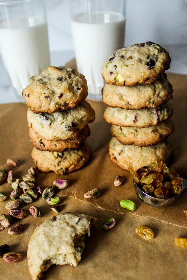 two stacks of cranberry raisin pistachio cookies on top of brown parchment paper with pistachios and golden raisins scattered in front and two glasses of milk behind them