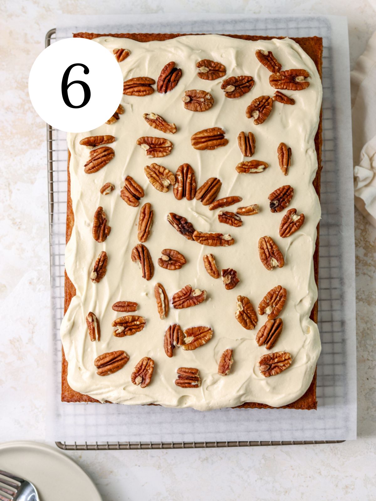 A rectangular cake topped with frosting and pecans on a wire rack.