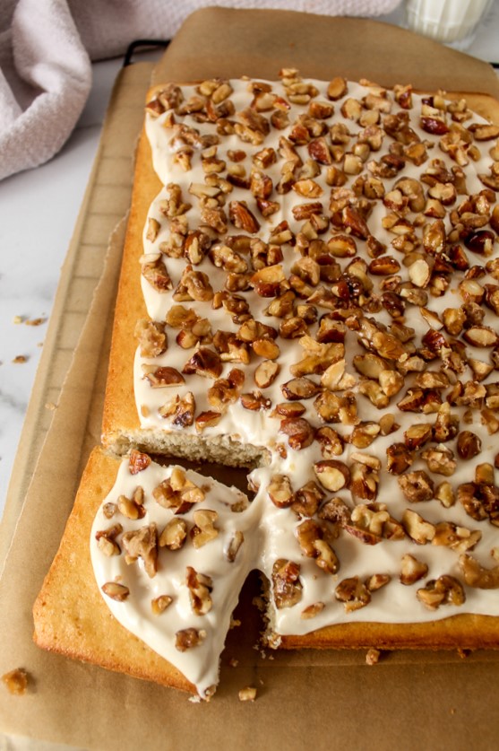 a large sheet cake topped with frosting and candied nuts on two sheets of parchment on top of a cooling rack with a light purple towel behind it and a glass of milk. 