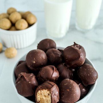 a white bowl filled with chocolate covered peanut butter balls. one balls has a bite in it and a white bowl of peanut butter balls behind it and two glasses of milk in the background