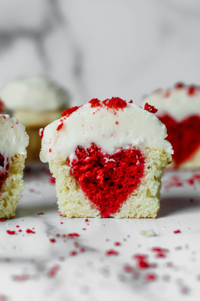 A white cupcake with a heart inside topped with buttercream and red velvet crumbs.
