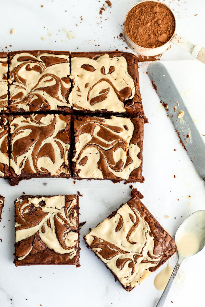 Fudgy, dense brownie squares topped with chocolate and tahini with cocoa, a knife and spoon next to them. 