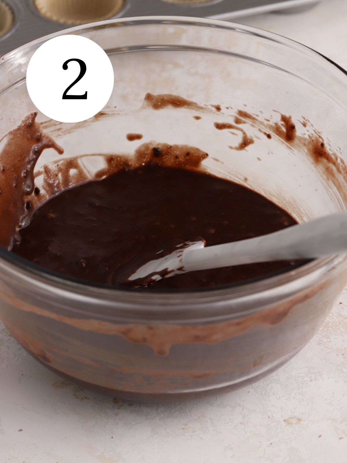 A glass mixing bowl with chocolate cupcake batter being stirred with a spatula.