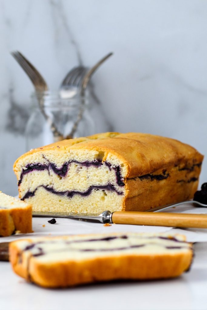 A delicious blueberry pound cake with homemade blueberry sauce swirled in it being sliced on a cutting board.