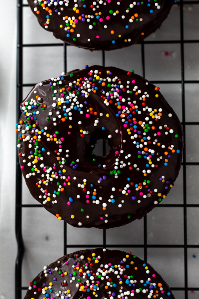 Chocolate donuts with chocolate ganache and rainbow sprinkles on a cooling rack.