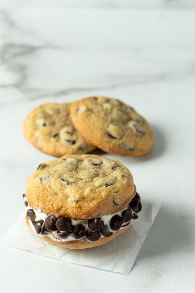 A thick, delicious chocolate chip ice cream sandwich with chocolate chip cookies behind it. 