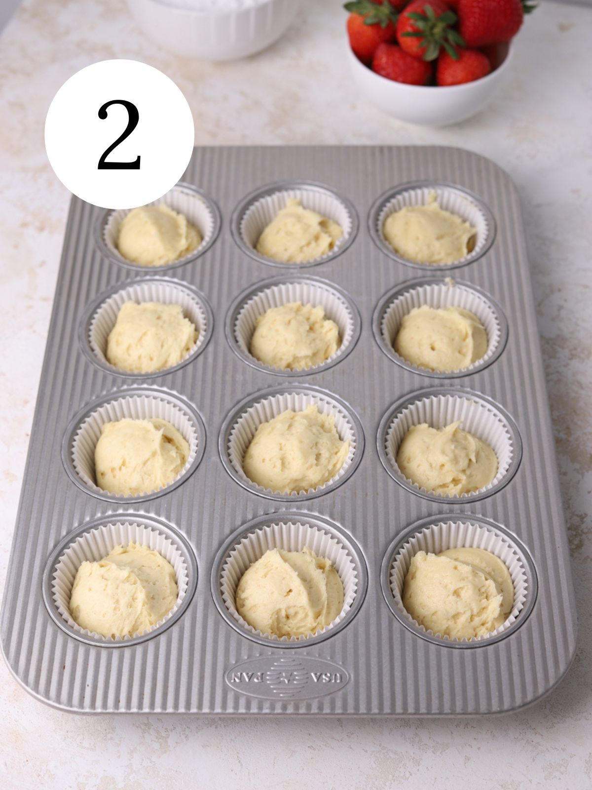 A muffin pan with liners and cupcake batter next to a bowl of strawberries.