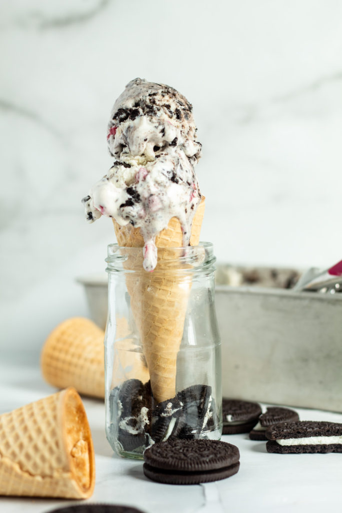 A cone filled with two scoops of creamy oreo ice cream with oreos and cones nearby.