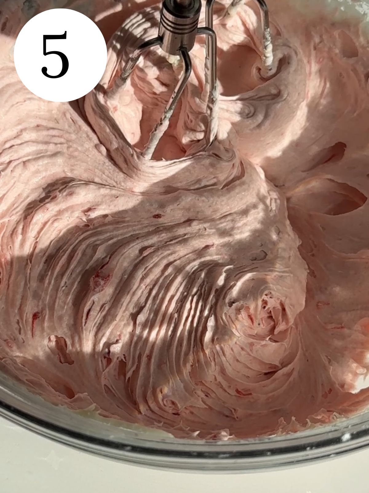 Strawberry frosting being mixed together with an electric mixer in a bowl.