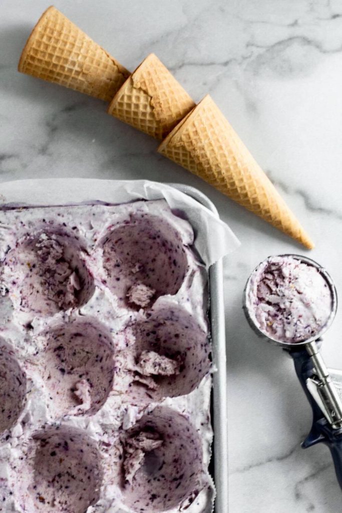 Berry ice cream in a pan with an ice cream scoop and cones next to it. 