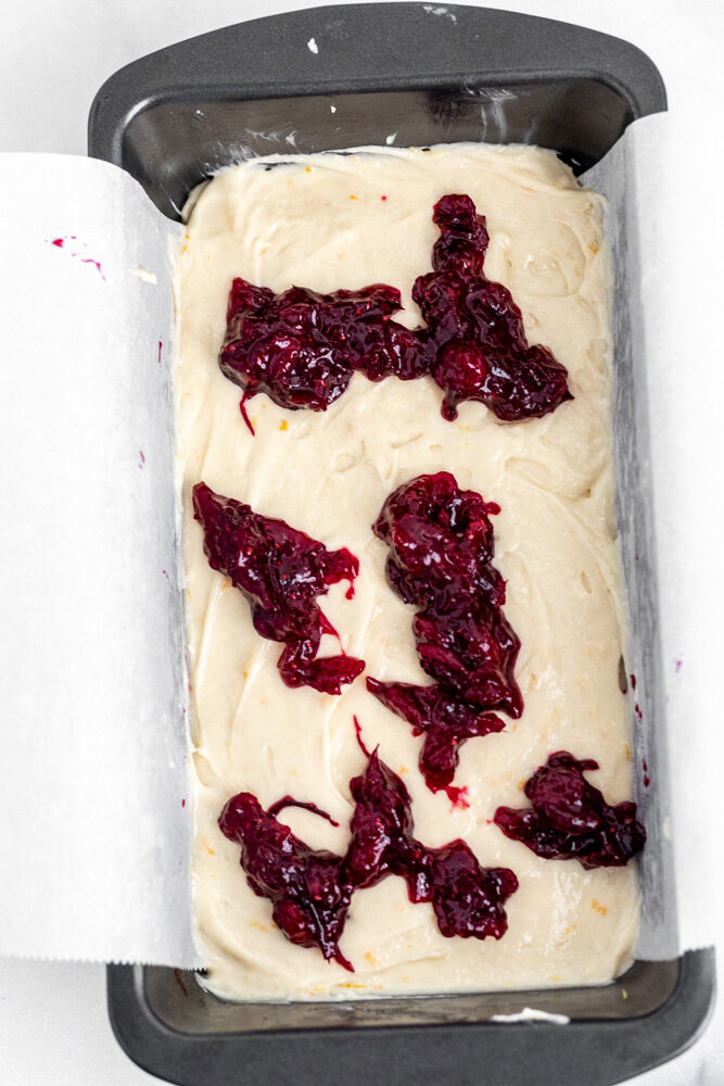 Pound cake batter in a pan lined with parchment paper topped with dollops of cranberry sauce. 