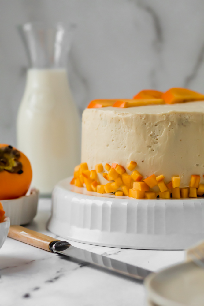 A close up of a persimmon layer cake with fresh persimmons on it next to a milk jug. 