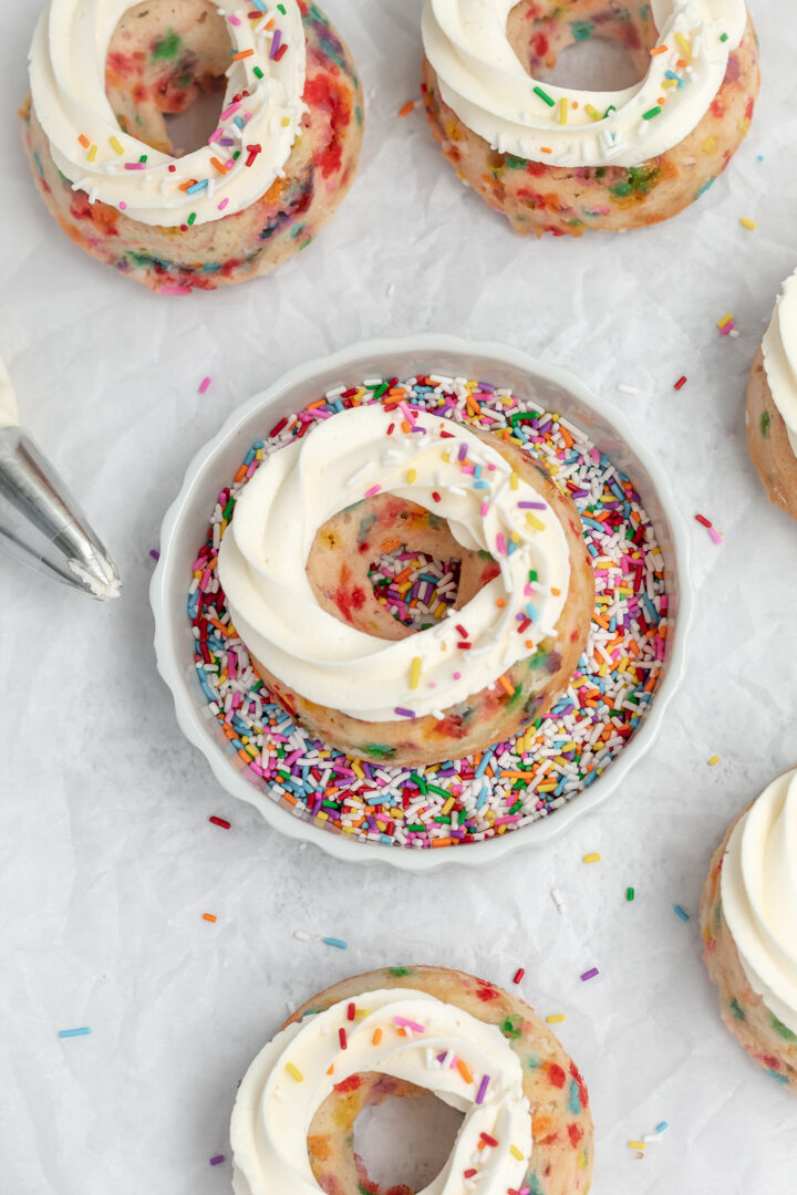A rainbow donut topped with a swirl of frosting in a bowl of sprinkles. 