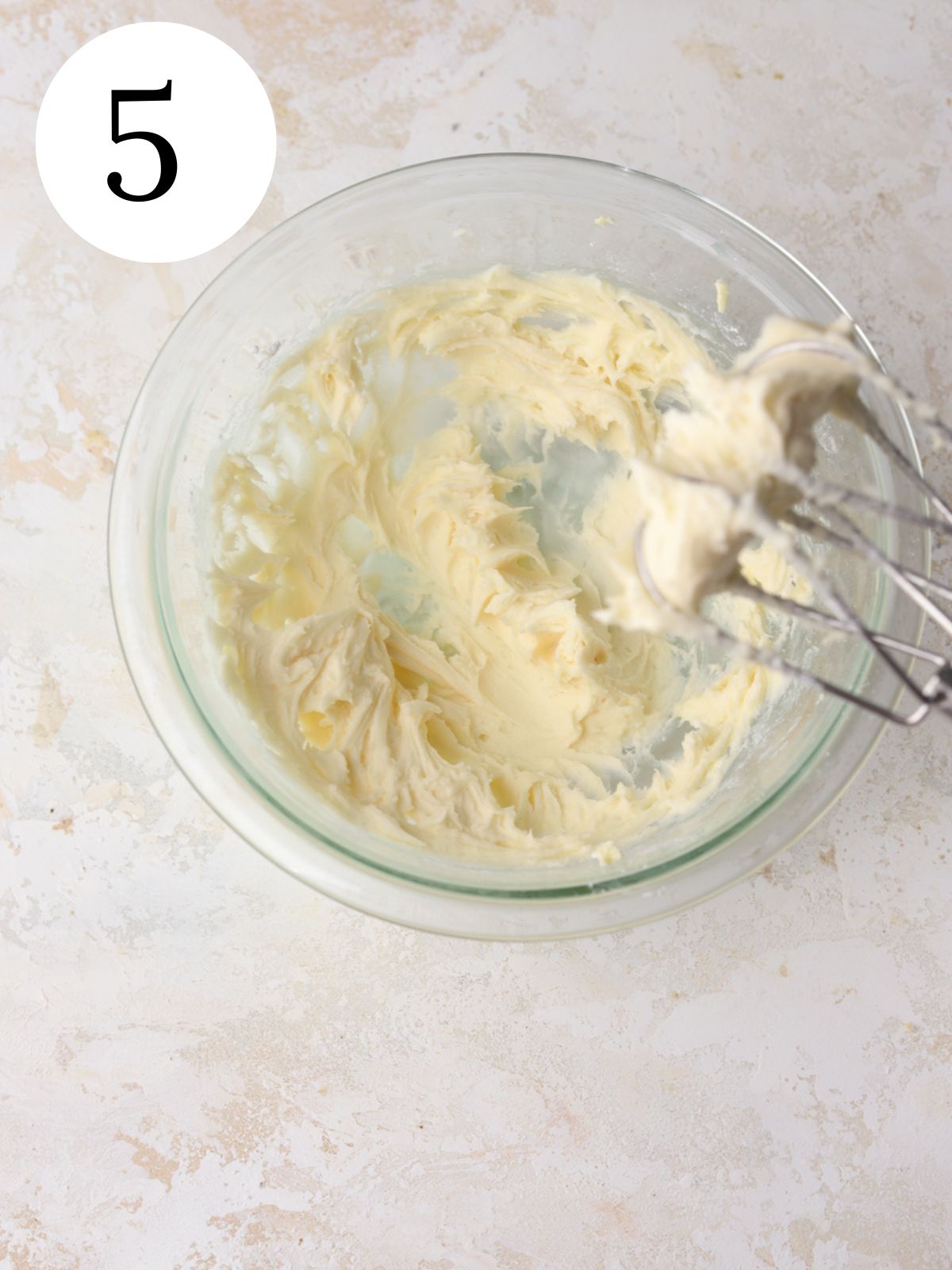A glass mixing bowl with frosting and a mixer.
