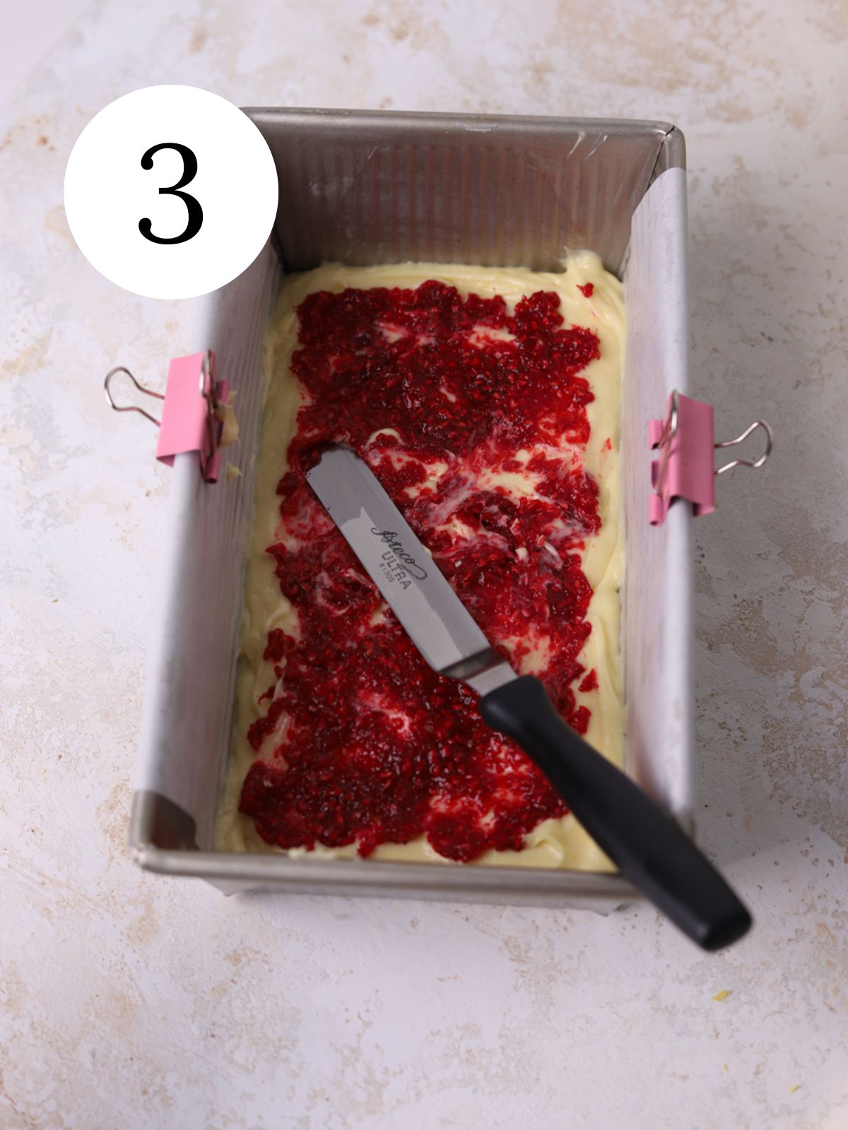 A loaf pan with raspberry jam being spread on cake batter with an offset spatula.