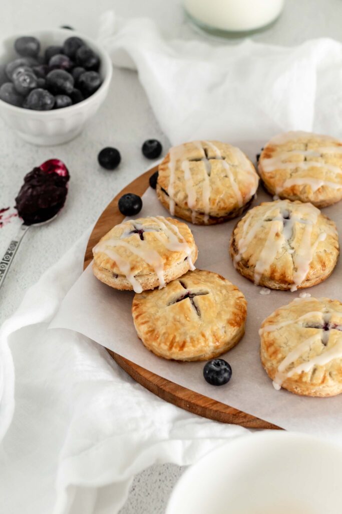 A batch of mini pies on parchment paper with blueberries scattered around them.