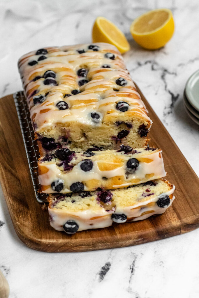Blueberry loaf cake topped with icing cut into slices on a wooden board.
