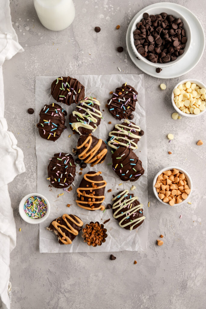 Chocolate covered eggs on parchment paper and bowls of chocolate chips and sprinkles. 