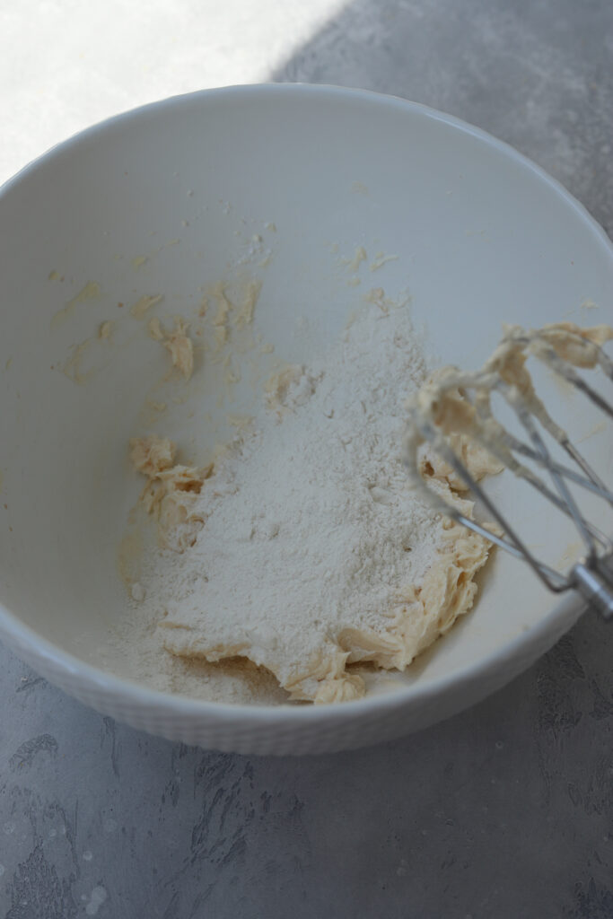 A large white bowl with flour on top of cookie dough and beaters next to the bowl.
