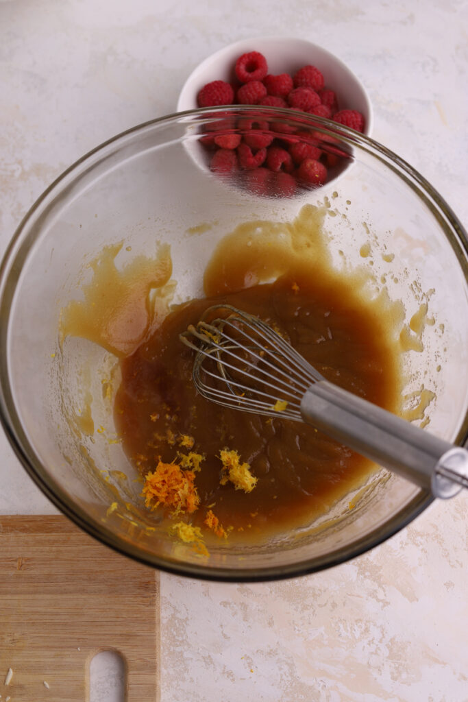 A glass bowl with wet ingredients, a whisk and orange zest in it. 