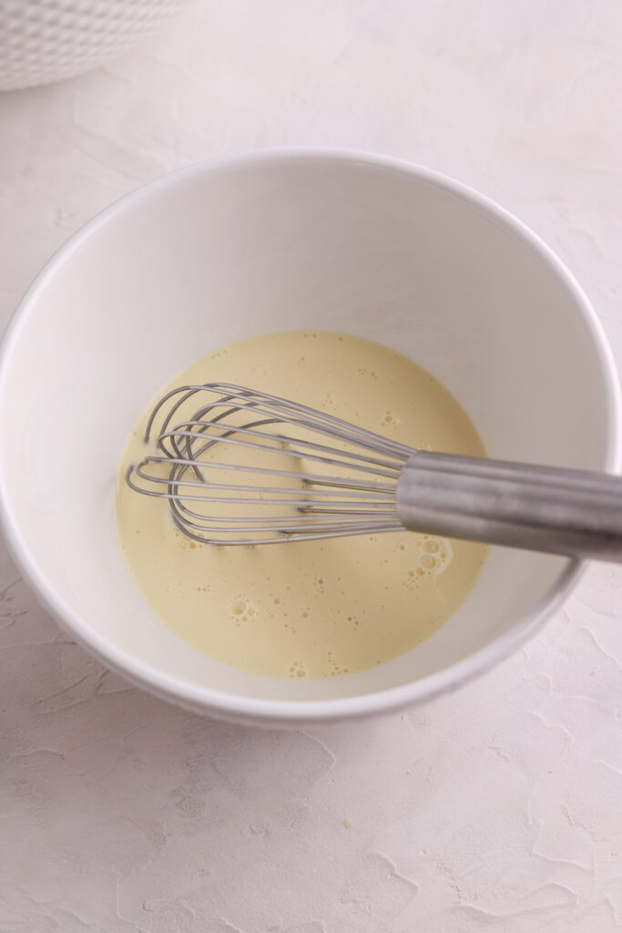 A white bowl with wet ingredients and a whisk in it.