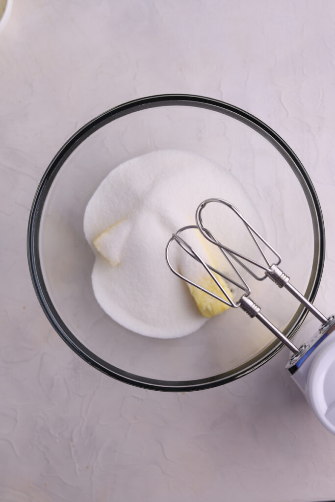 A glass bowl with sugar and butter in it and a mixer. 