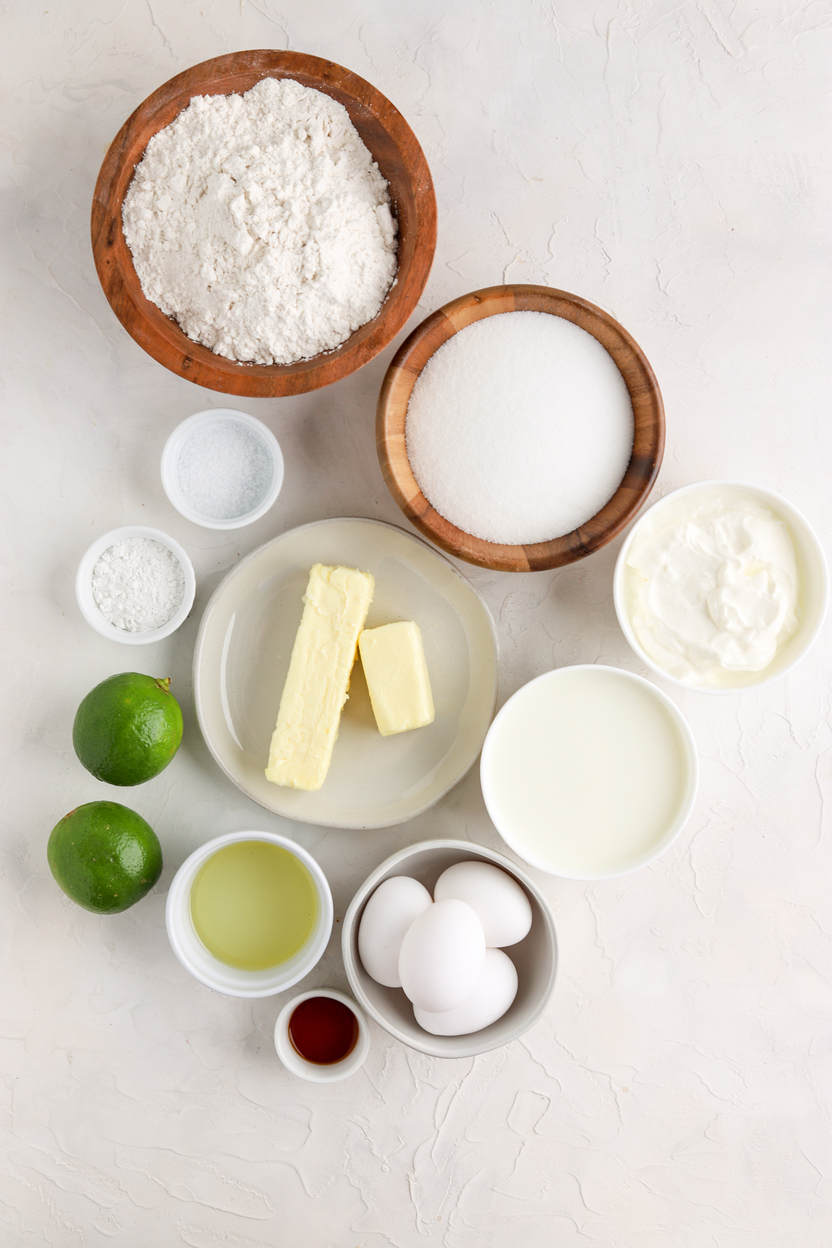 Flour and sugar in bowls, butter on a plate, eggs in a bowl and two limes. 
