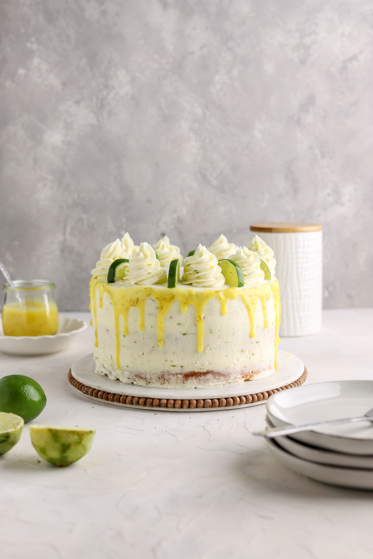 Lime cake on a cake stand with limes and plates next to it. 