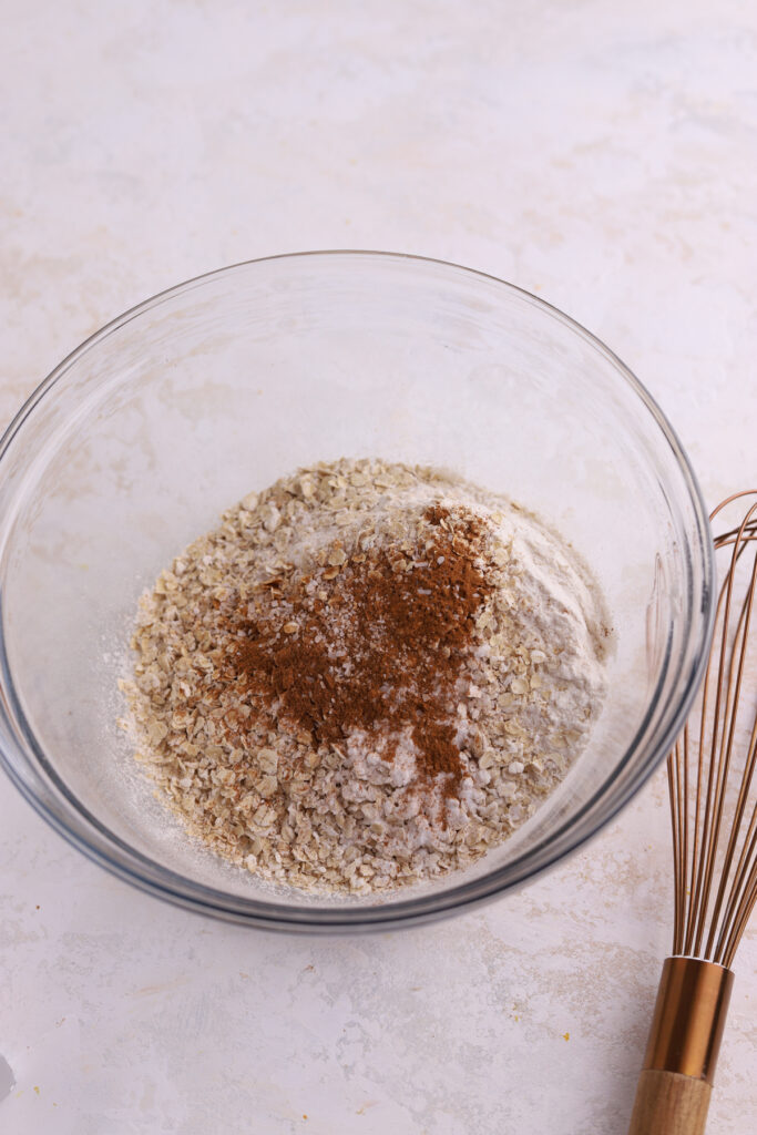 A mixing bowl with oatmeal, flour and cinnamon in it and a gold whisk. 