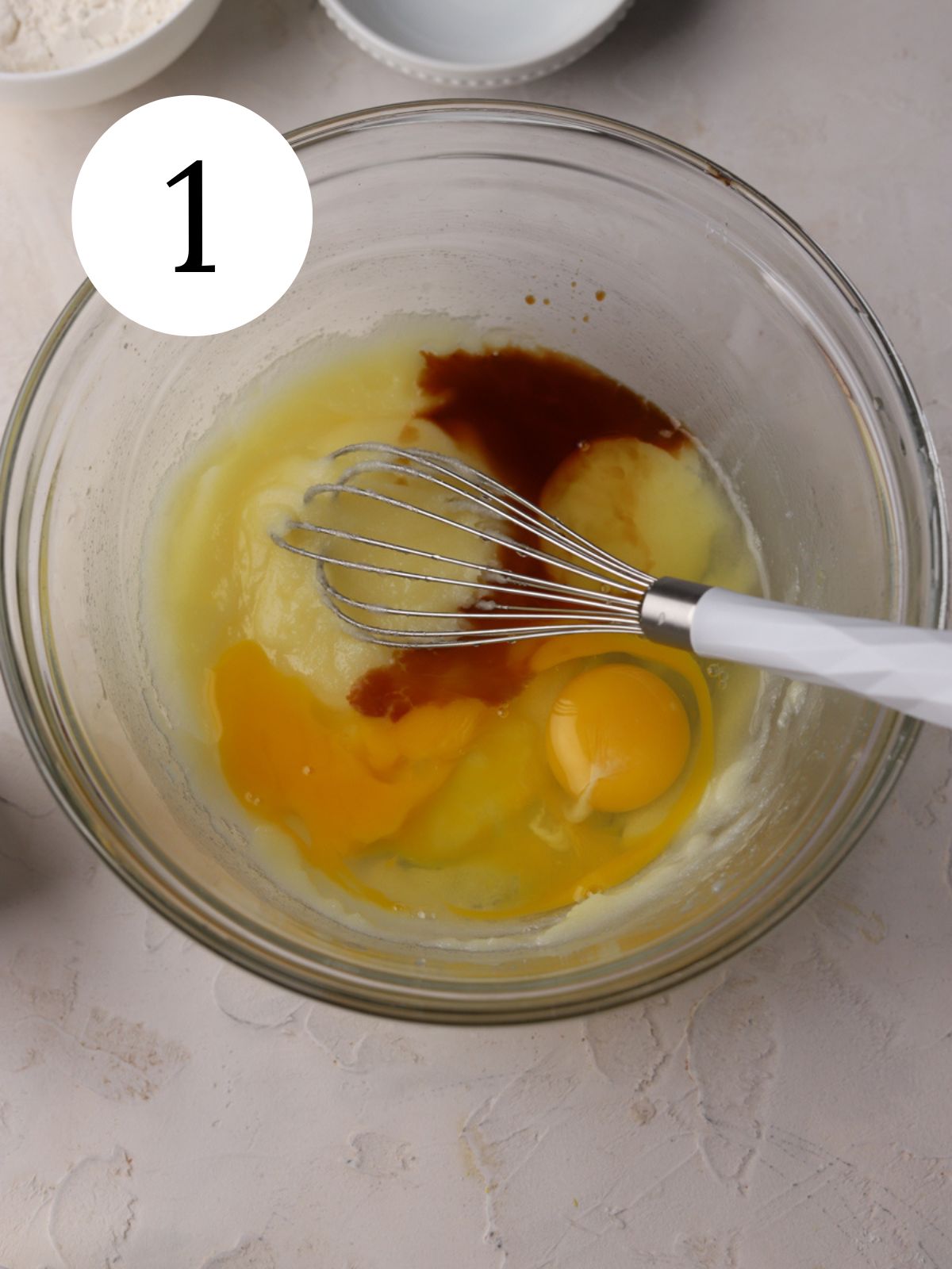 A glass mixing bowl with eggs, sugar and vanilla and a whisk.