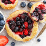 Mini blueberry strawberry pies and fresh blueberries and strawberries on parchment paper.