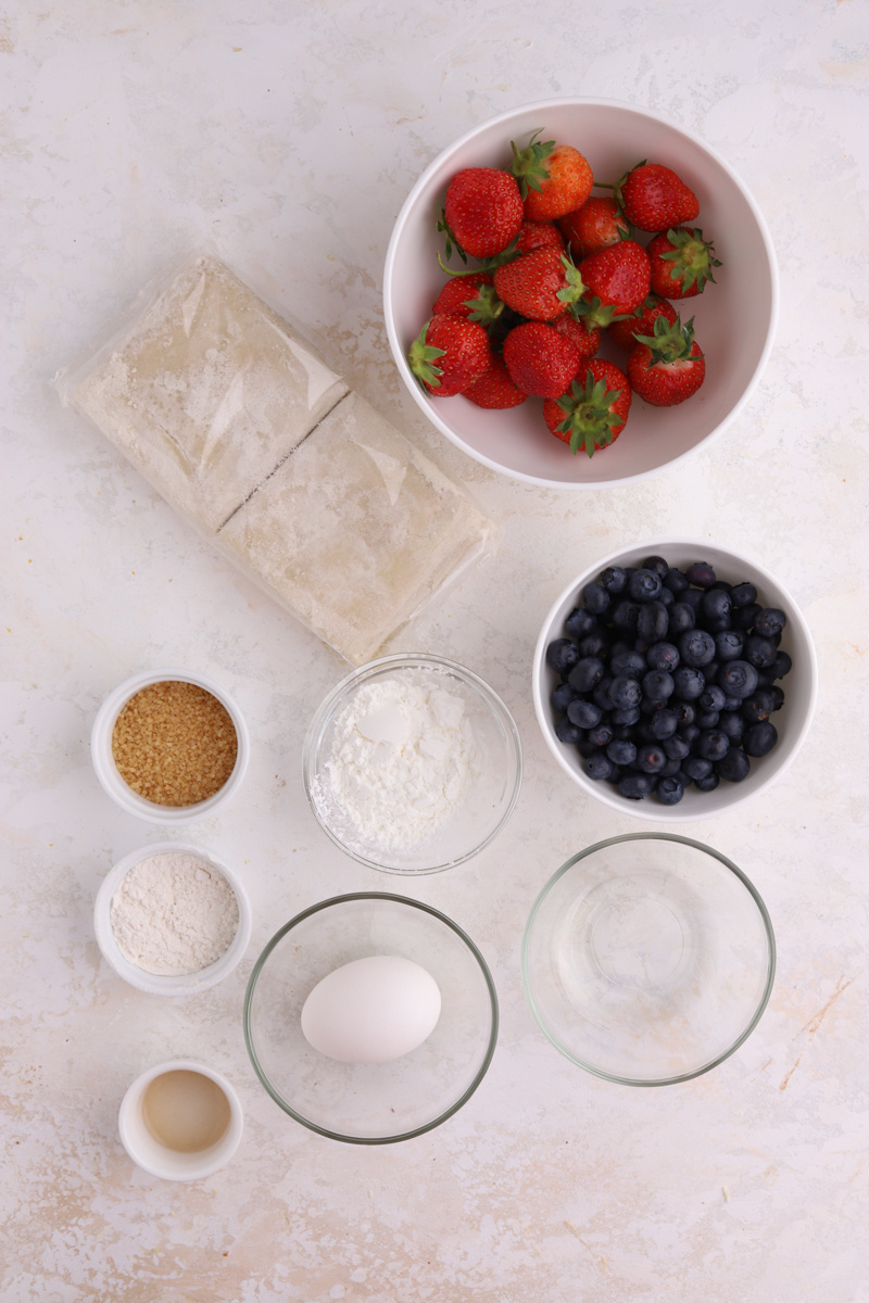 Bowls filled with strawberries, blueberries, an egg and sugar and a package of puff pastry.