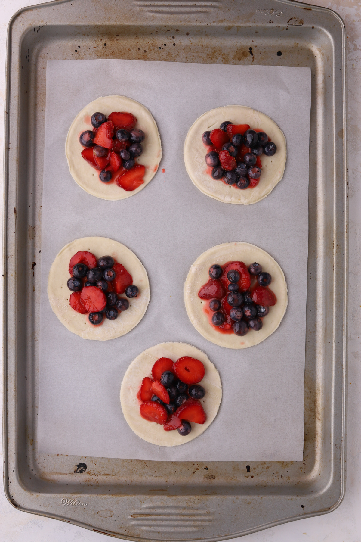 A baking tray lined with parchment paper with blueberries and strawberries on small pie crusts.