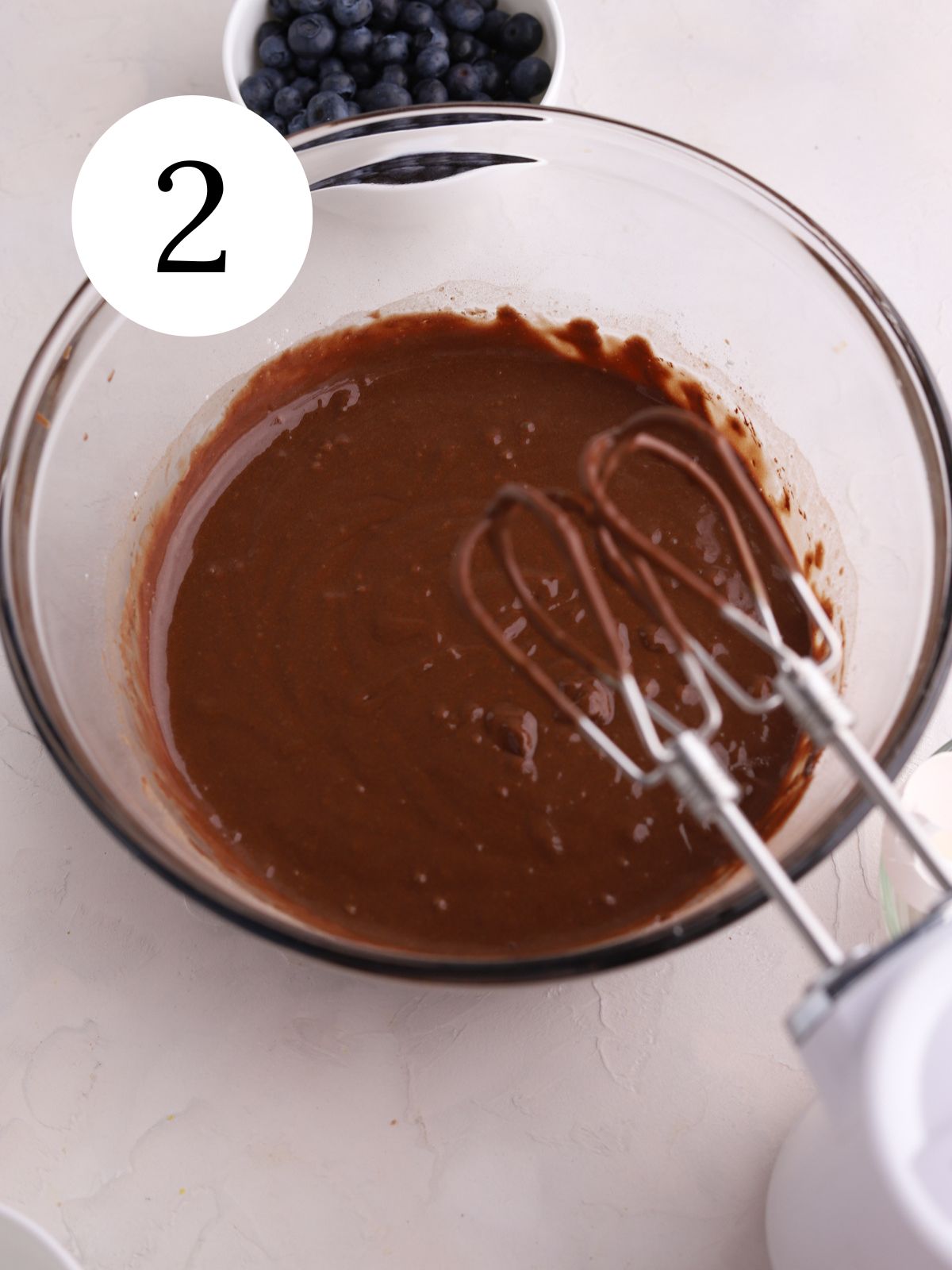 Chocolate cake batter in a bowl with a mixer and a bowl of blueberries.