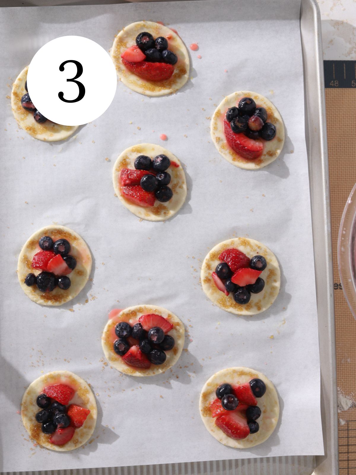 A baking tray lined with parchment paper with blueberries and strawberries on small pie crusts.